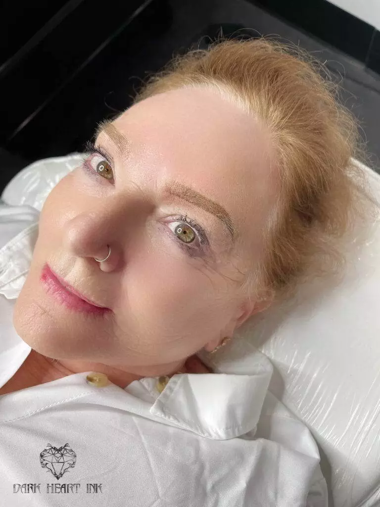 A woman with closed eyes receiving permanent eyebrow makeup on a bed.