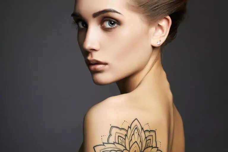 A person with a floral tattoo on their back, inked by a qualified cosmetic tattoo artist in San Diego, looks over their shoulder, displaying a neutral expression and wearing small stud earrings.