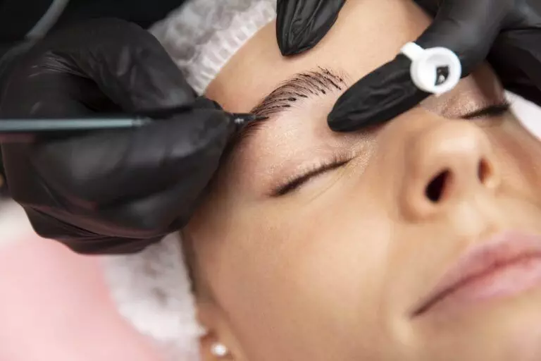 In San Diego, a person receives an eyebrow tattoo procedure with black gloved hands using a tool to create fine strokes on their permanent eyebrows while they lay down with closed eyes.