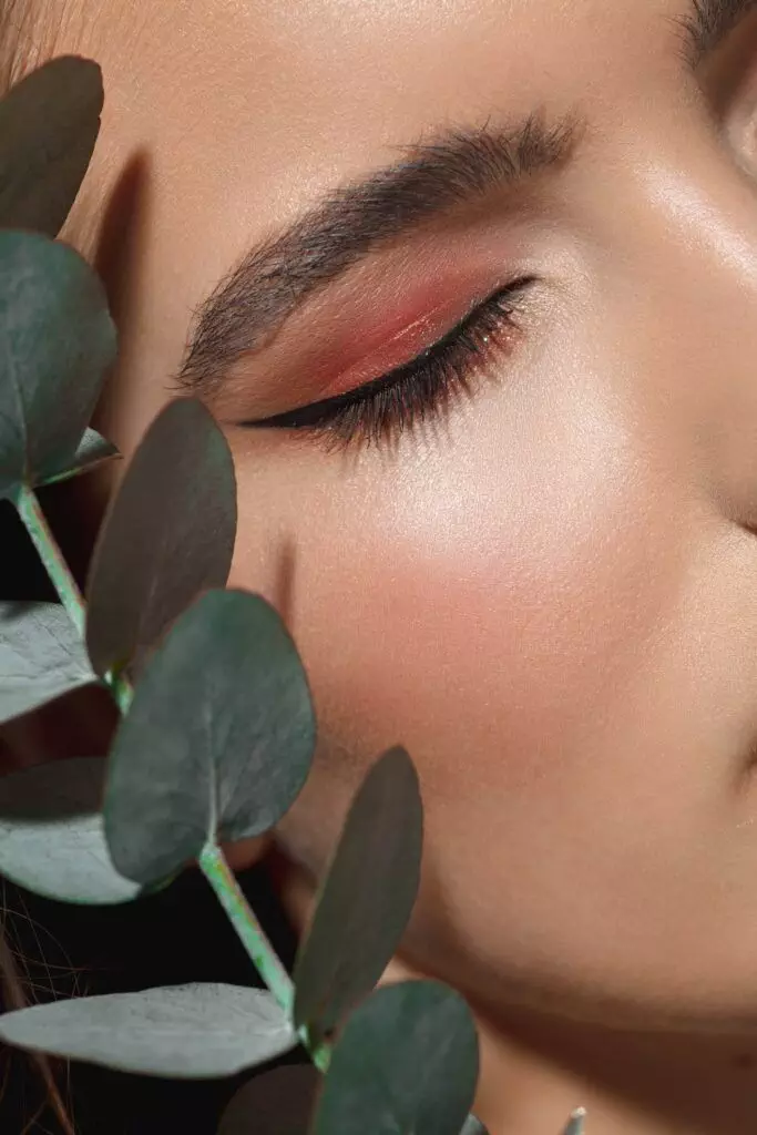 Close-up of a person's face with closed eyes and pink eye shadow, framed by green eucalyptus leaves, showcasing the elegance of permanent eyeliner.