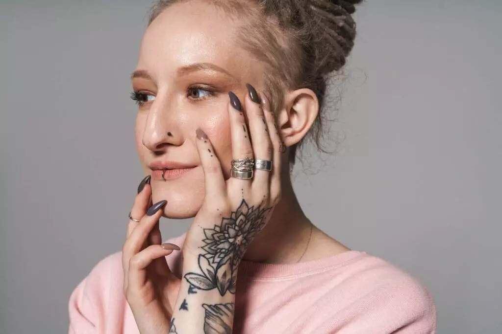 A person with tattoos on their hand and long, painted nails poses with their hands touching their face, wearing rings and a light pink top, showcasing the artistry of Dark Heart Ink.