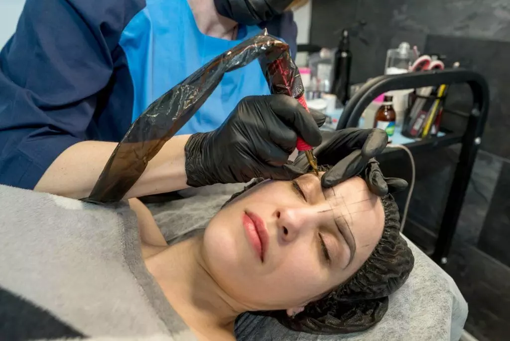 A person is receiving cosmetic tattooing from a technician wearing gloves and a face mask. The person lies on a treatment bed with eyes closed, fully aware that proper aftercare is essential for optimal healing.