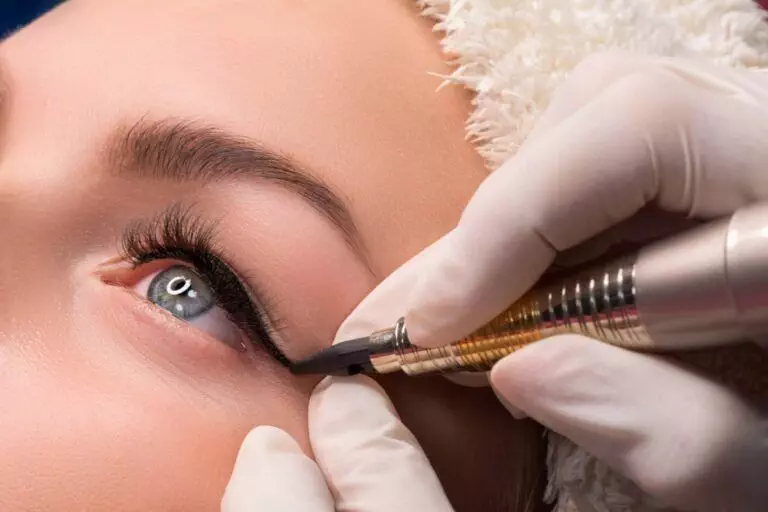 Close-up of a person receiving a cosmetic tattoo at Dark Heart Ink, with gloved hands applying black eyeliner using a tattoo machine on the upper eyelid, expertly transitioning from Classic to Dramatic permanent eyeliner styles.