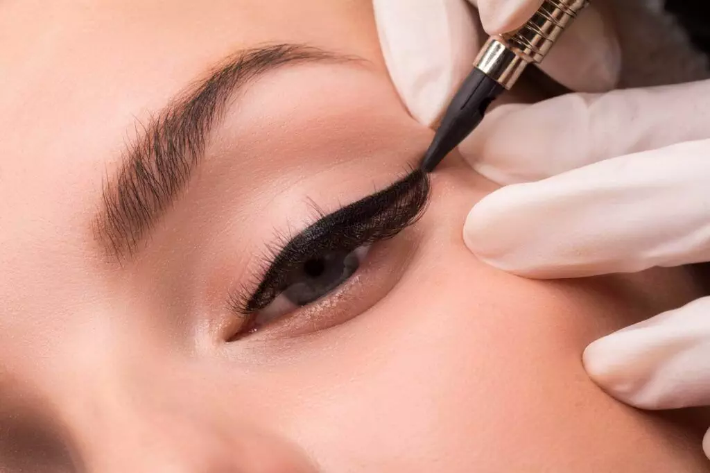 A close-up shows a cosmetician's gloved hand applying permanent eyeliner on a person's eyelid using a precision tool, ensuring no smudging or need for reapplication.