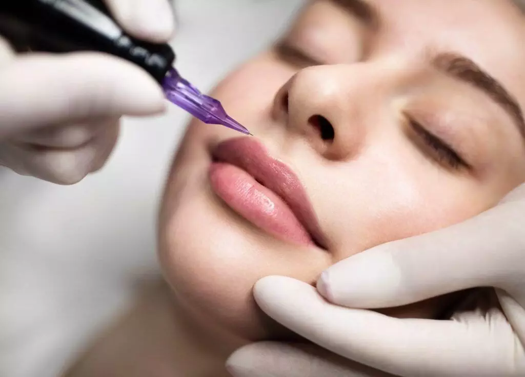 A close-up of a person receiving a cosmetic lip tattoo procedure, often referred to as lip blushing. Hands in gloves hold a tattoo machine with a purple needle near the person's lips. This precise and meticulous beauty treatment is becoming increasingly popular in San Diego.