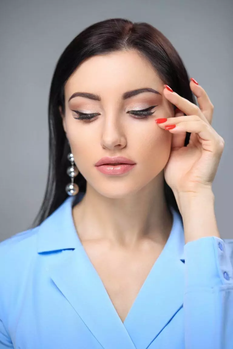 A woman with long dark hair, adorned with pearl earrings and a blue blazer, has her eyes closed and touches her temple with a hand, showcasing her perfectly applied permanent eyeliner.