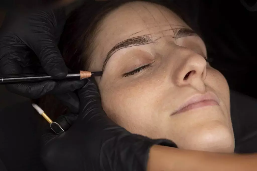 A cosmetologist with gloved hands uses a pencil to draw an eyebrow outline on a woman's face as she keeps her eyes closed, preparing for natural-looking eyebrow microshading.