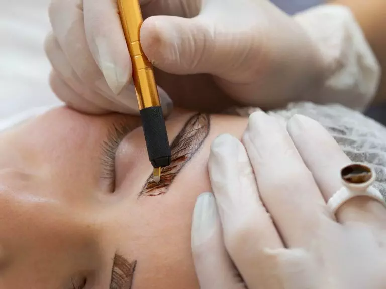 A professional is performing a cosmetic tattooing procedure on a person's eyebrows using a specialized tool, while wearing gloves.