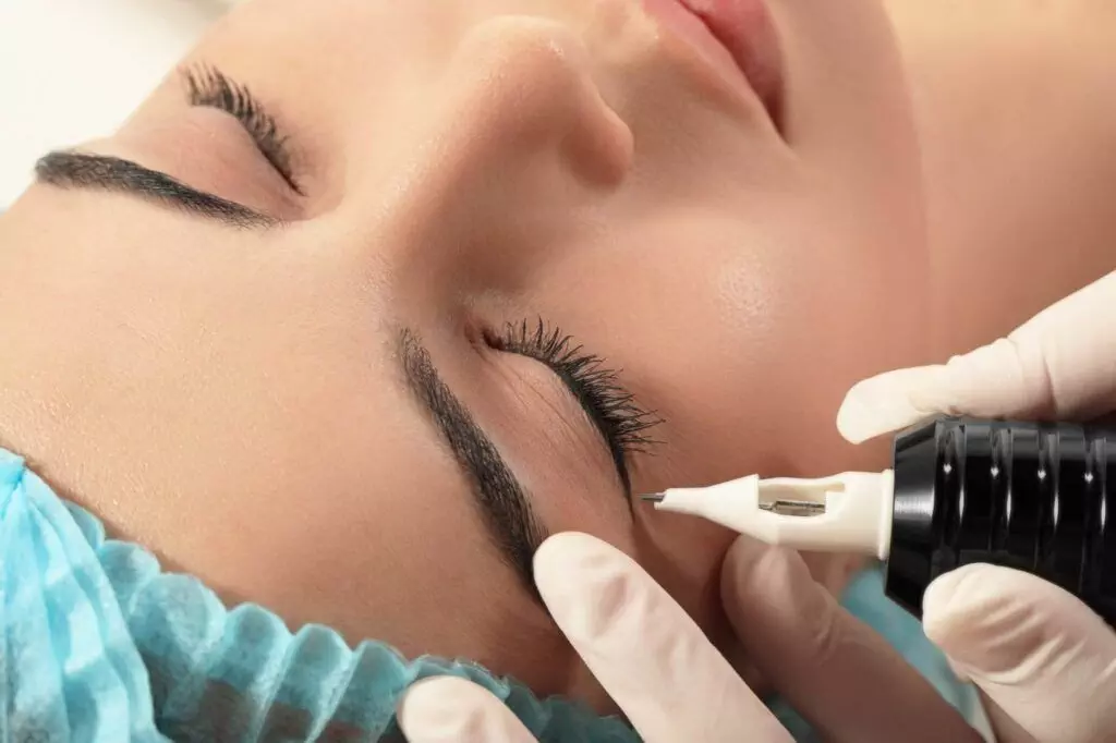 A person is receiving an eyebrow tattoo procedure at Dark Heart Ink, with a close-up view showing hands in gloves using a tattoo machine on the person's eyebrow area. The individual, who is also considering different eyeliner styles for their next session, is wearing a blue head covering.