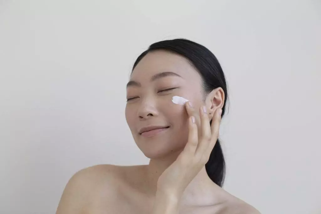 A person applies a small amount of cream to their cheek with closed eyes and a serene expression, showcasing trendy beauty enhanced by natural-looking beauty marks.