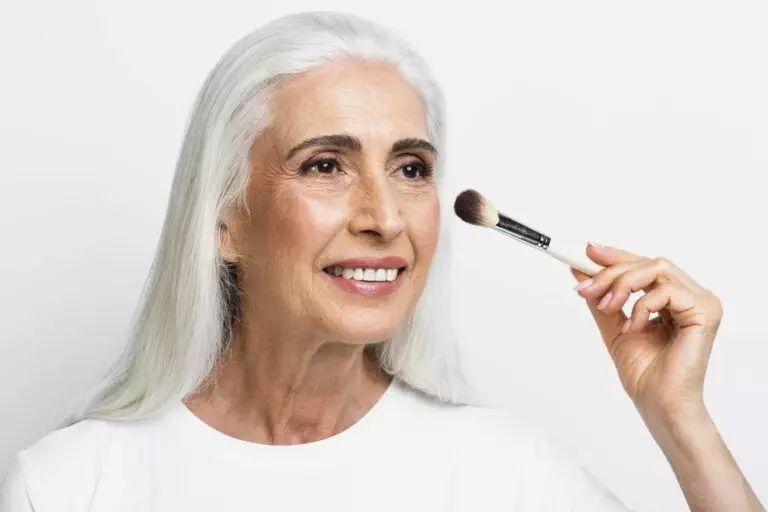 An older adult with long gray hair holds a makeup brush to her cheek, highlighting her mature skin and smiling against a white background.