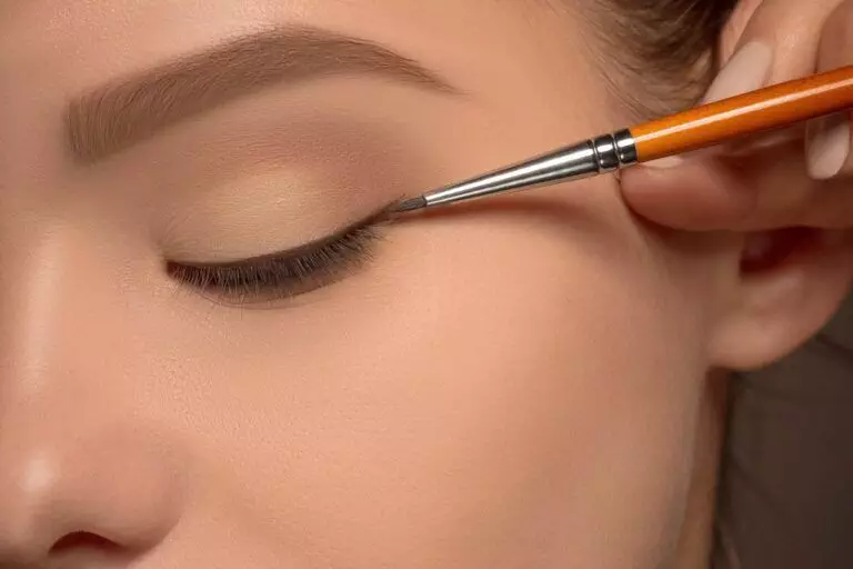 A close-up of a person applying black eyeliner with a brush to their closed eyelid, reflecting the effortless beauty often achieved through cosmetic winged eyeliner tattoos.